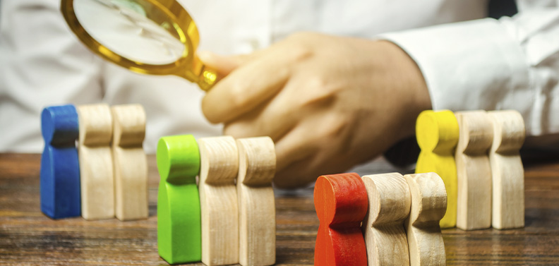 homem colocando uma lupa sobre vários bonequinhos de madeira em grupos separados por cor, indicando uma estratégia de marketing personalizada