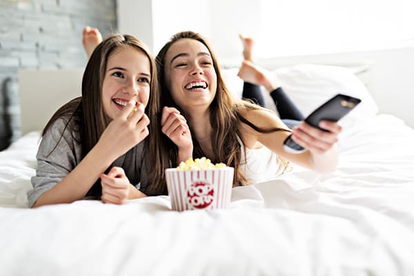 Duas adolescentes assistindo televisão deitadas na cama e comendo pipoca.