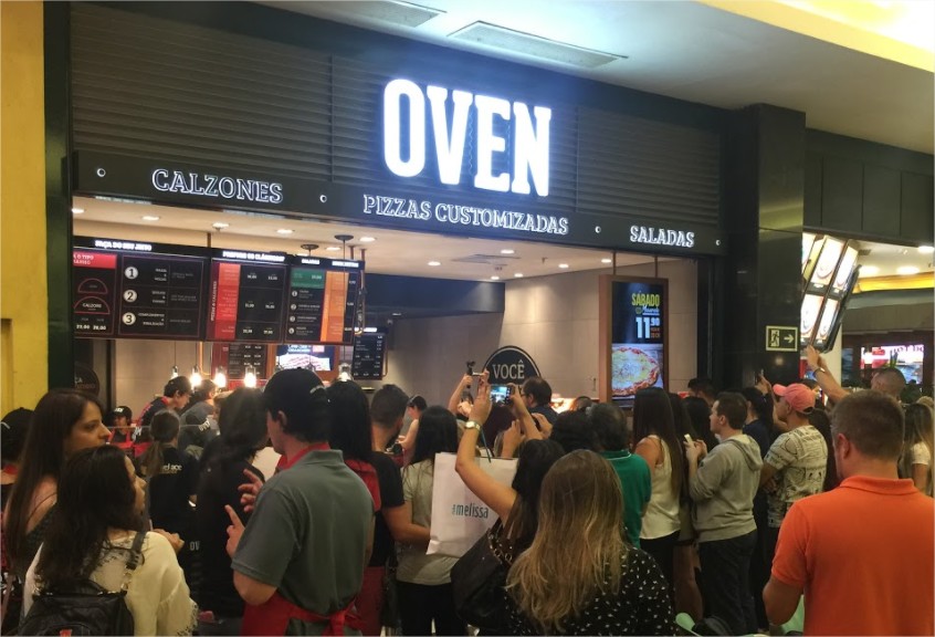Fachada da Oven Pizza em uma praça de alimentação lotada de clientes.