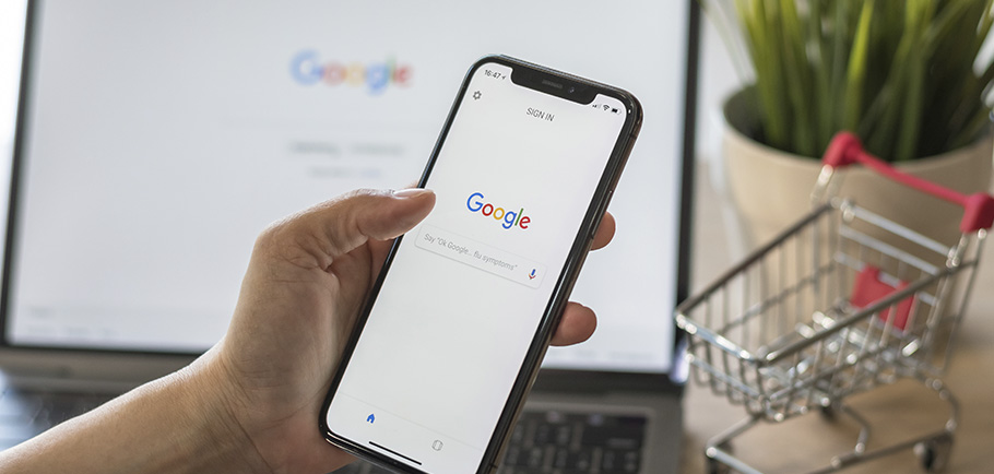 Pessoa segurando um celular com a tela de pesquisa do Google aberta, ao fundo está um carrinho de mercado indicando uma compra online.